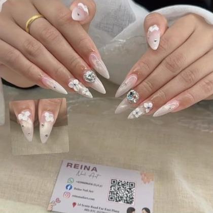 Almond nails with raised white flowers and small crystals, photographed on linen beside a Reina Nail Art card